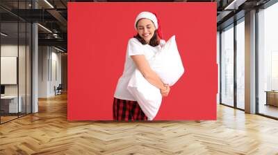 Beautiful young woman in pajamas and Santa hat with pillow on red background Wall mural