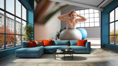 Beautiful young woman in sportswear exercising on fitness ball at gym Wall mural