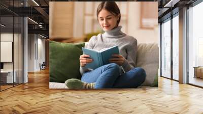 Beautiful young woman reading book on sofa at home Wall mural