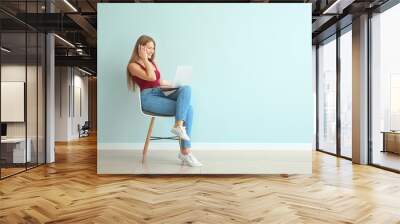 Beautiful young woman with laptop sitting on chair against color wall Wall mural