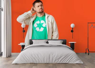 Confused young man in t-shirt with recycling logo scratching head on orange background. Ecology concept Wall mural