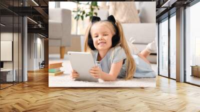 Cute little girl in headphones listening to audiobook with modern tablet computer while lying on carpet in living room Wall mural