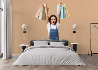 Fashionable young Asian woman in stylish denim overalls with shopping bags sitting against brown background Wall mural