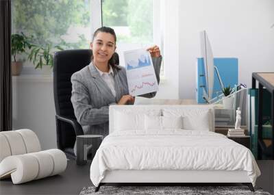 Female office worker showing paper sheet with graphs and sitting on armchair at workplace in light office Wall mural