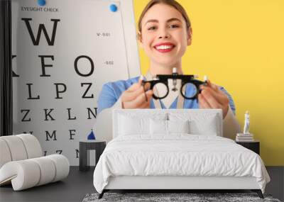 Female ophthalmologist with trial frame and eye test chart on yellow background, closeup Wall mural