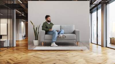 Handsome bearded man using laptop on grey sofa near light wall Wall mural