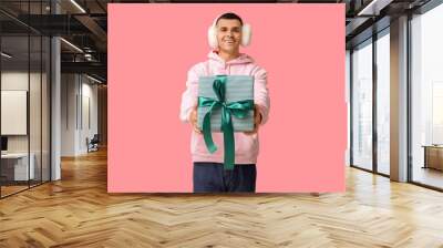 Handsome young man in warm fluffy earmuffs holding Christmas gift on pink background Wall mural