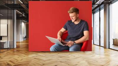 Handsome young man with laptop sitting on chair against color background Wall mural