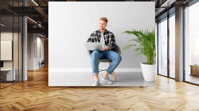 handsome young man with laptop sitting on chair near light wall Wall mural