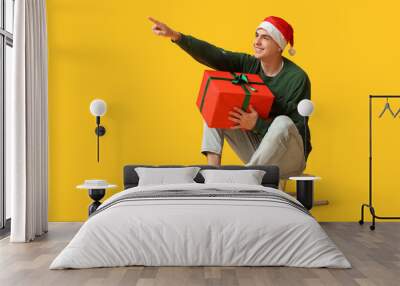 Happy young man in Christmas sweater and Santa hat with gift box on sledge against yellow background Wall mural