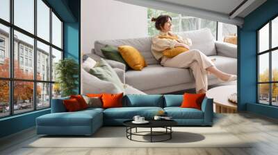 Happy young woman sitting on grey sofa in interior of light living room Wall mural