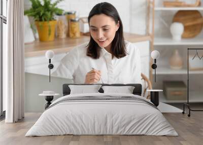 Happy young woman with bowl of tasty yoghurt and laptop at table in kitchen Wall mural