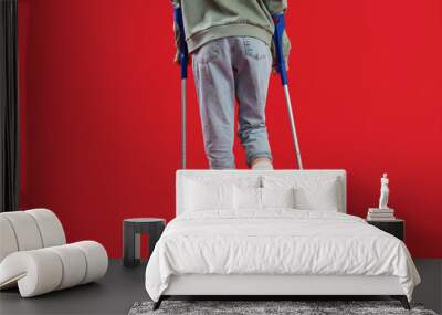 Injured young woman after accident with broken foot and crutches on red background, back view Wall mural