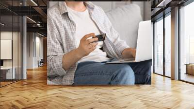 Man with credit card and modern laptop sitting on sofa while shopping online, closeup Wall mural