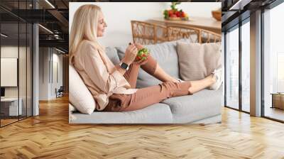 Mature woman eating vegetable salad on sofa at home Wall mural