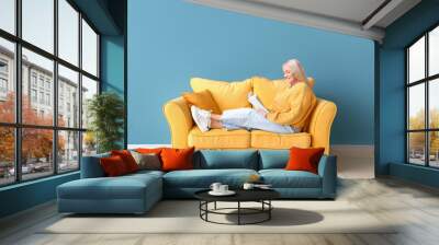 Mature woman reading book while sitting on sofa near blue wall Wall mural