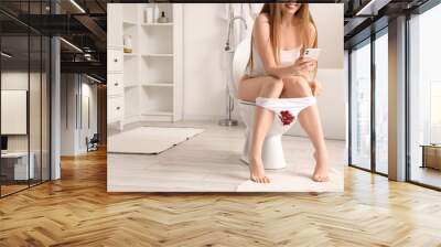 Menstruating woman with stained panties using mobile phone on toilet bowl in restroom. Free bleeding concept Wall mural