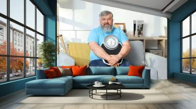 Overweight happy mature man with scales sitting on sofa at home. Weight loss concept Wall mural