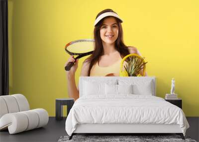 Portrait of pretty young woman in swimsuit with pineapple, headphones and tennis racquet on yellow background Wall mural