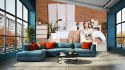 Pretty young woman with fashion magazine sitting near bed in light bedroom Wall mural