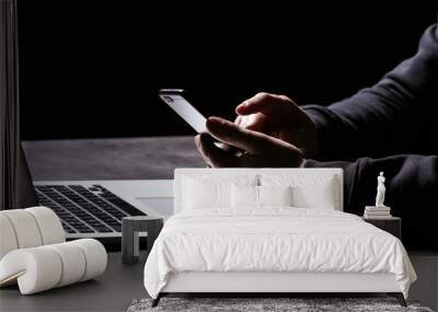 Professional hacker with laptop and mobile phone sitting at table on dark background, closeup Wall mural