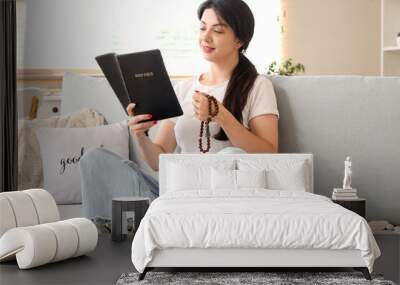 Religious young woman with praying beads reading Holy Bible on sofa at home Wall mural