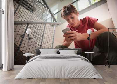 Sad teenage boy with mobile phone sitting on stairs. Bullying at school Wall mural