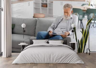 Senior bearded man reading magazine on sofa in kitchen Wall mural