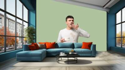 Sleepy young man in dirty t-shirt with saucer and cup of coffee on green background Wall mural