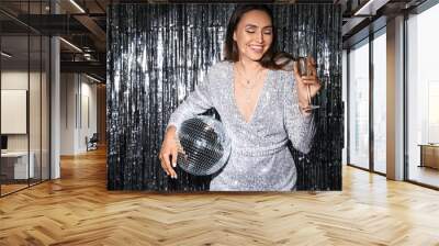 Smiling fashionable woman in sparkling dress with disco ball and glass of champagne against silver tinsel Wall mural