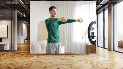 Sporty happy male blogger with dumbbells recording video at home Wall mural