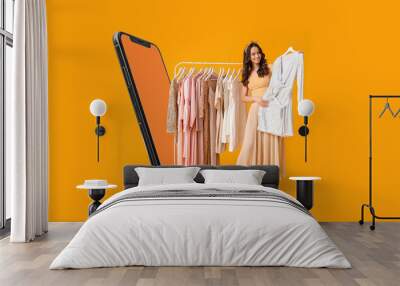 Stylish young woman with stylish clothes on hanger and modern mobile phone on yellow background Wall mural