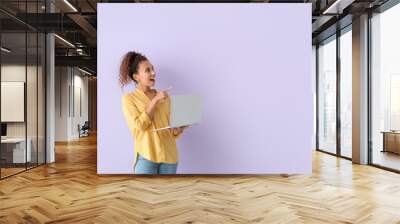 Surprised young African-American woman with laptop pointing at something on lilac background Wall mural