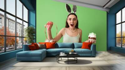 Surprised young woman with tasty Easter cake and eggs in basket on green background Wall mural