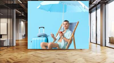 Teenage boy with soda using mobile phone in deck chair on blue background Wall mural