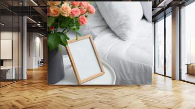 Vase with bouquet of beautiful fresh roses and blank frame on table in bedroom, closeup Wall mural