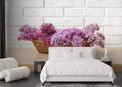 Wicker basket with beautiful lilac flowers on wooden table near light brick wall, closeup Wall mural