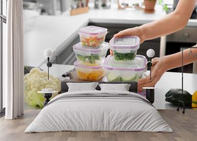 Woman holding stack of plastic containers with fresh vegetables for freezing at table in kitchen Wall mural