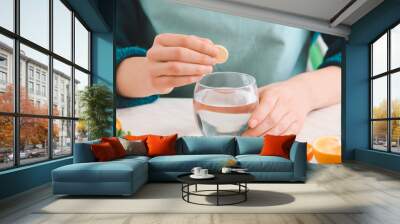 Woman putting ascorbic acid pill in glass of water on table in kitchen Wall mural