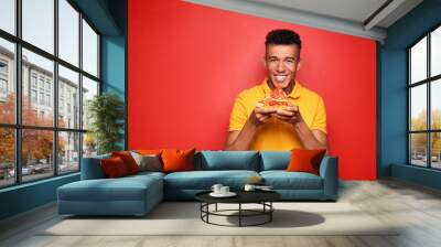 Young African-American man eating slice of delicious pizza on color background Wall mural