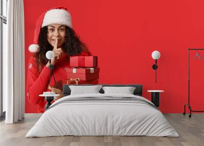 Young African-American woman in Christmas sweater and with gifts showing silent gesture on red background with space for text Wall mural