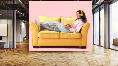 Young African-American woman with headphones using laptop on yellow sofa against pink background Wall mural