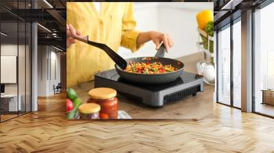 Young Asian woman cooking different vegetables in frying pan on portable electric stove at kitchen Wall mural