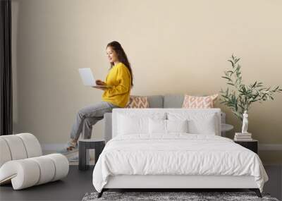 Young Asian woman using laptop on grey sofa near beige wall Wall mural