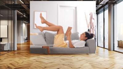 Young barefoot woman reading book on sofa at home Wall mural