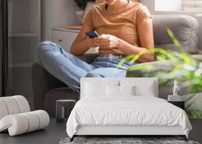 Young barefoot woman using mobile phone in armchair at home Wall mural