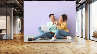 Young couple with laptop giving each other high-five near lilac wall Wall mural