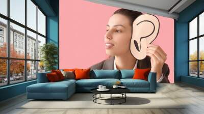 Young gossip businesswoman with paper ear trying to hear something on pink background, closeup Wall mural