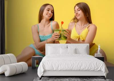 Young happy women in swimsuits with glasses of mojito sitting on inflatable mattress against yellow background Wall mural