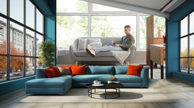 Young male freelancer with laptop lying on sofa in modern home office Wall mural